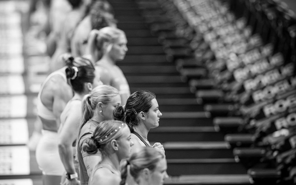 Female athletes standing in a line on the competition floor before an event, eyes forward, with rows of bumper plates behind them