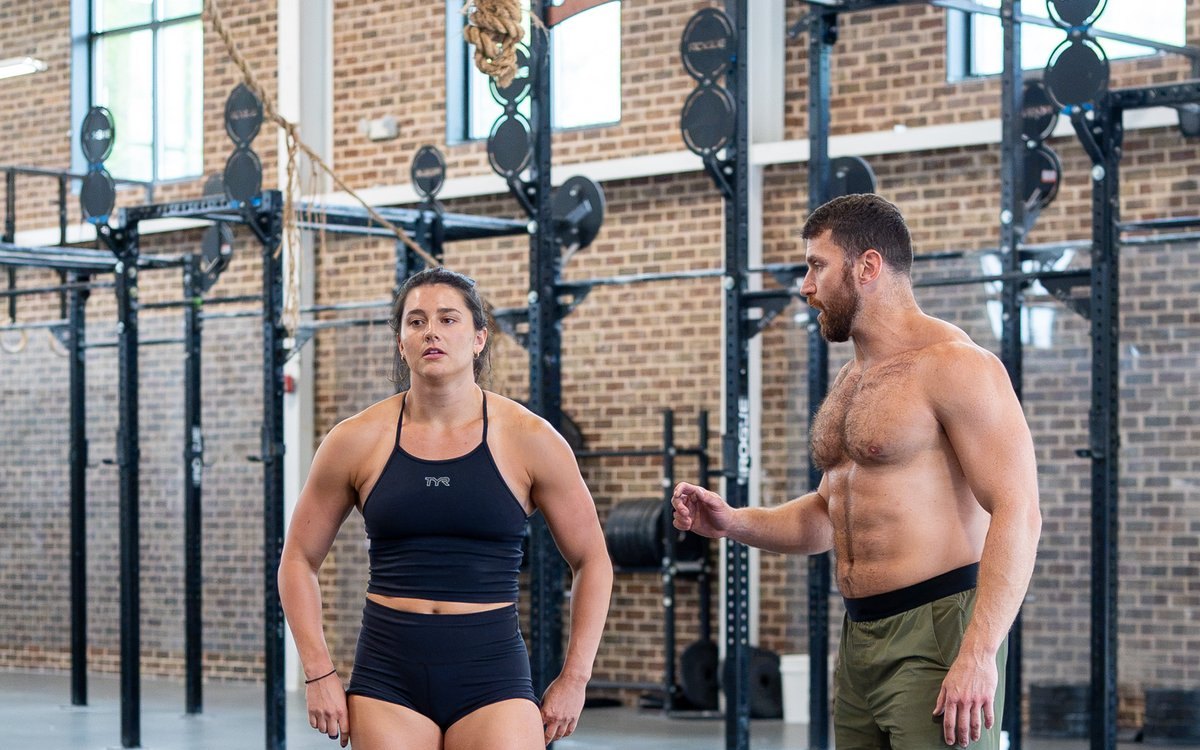 A coach talking an athlete through a cue between the rigs in a brick-walled gym