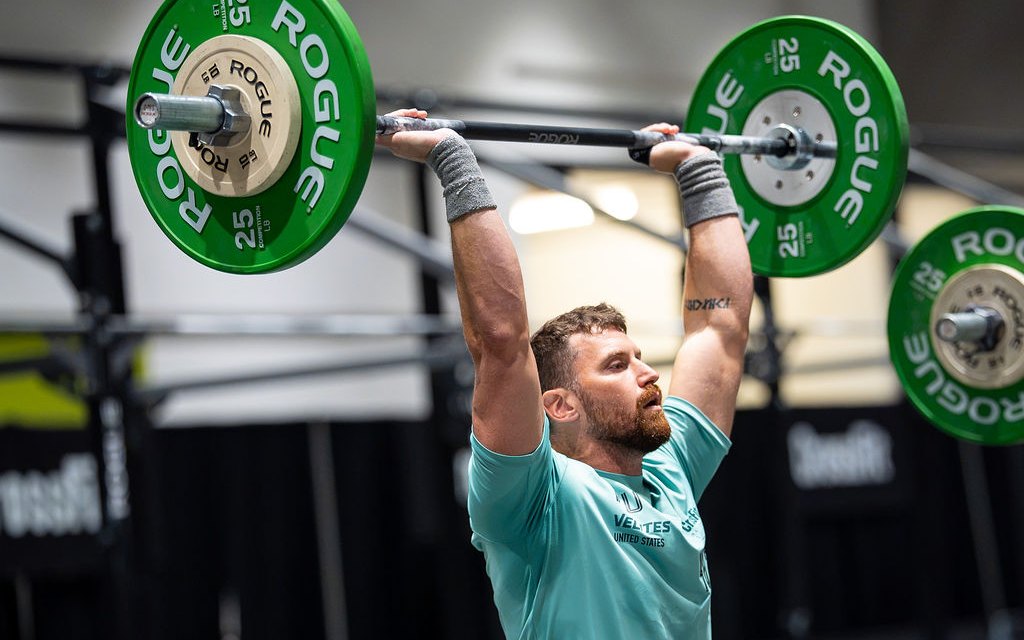 Kyle locking out a barbell overhead at the top of a thruster in a competition warm-up area