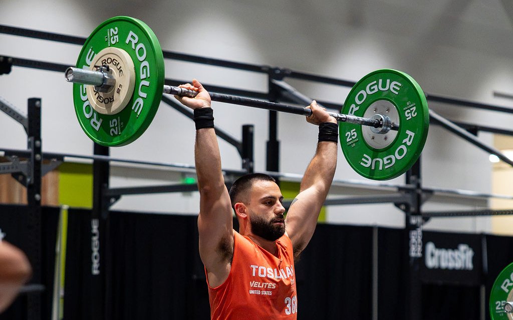 An athlete locking out a barbell overhead at the finish of a thruster at the 2026 CrossFit Games